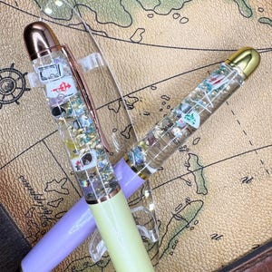 May include: Two clear pens with decorative elements inside, including playing card suits and glitter. One pen has a lavender barrel and rose gold accents, the other has a light green barrel and gold accents. Both pens have clear pen clips.