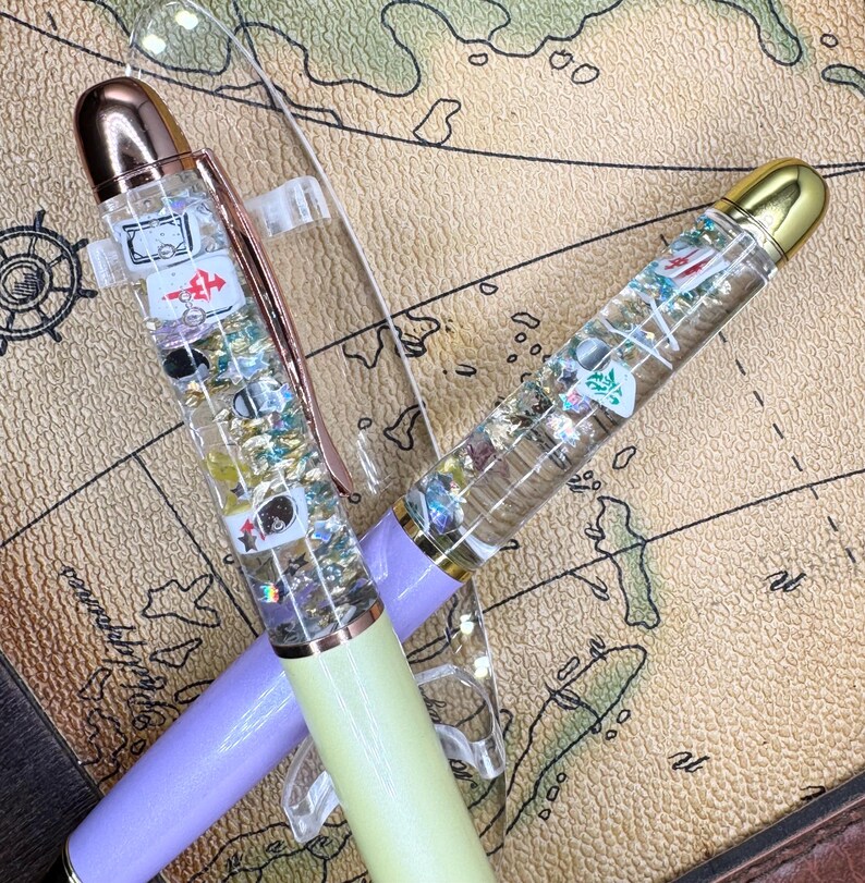 May include: Two clear pens with decorative elements inside, including playing card suits and glitter. One pen has a lavender barrel and rose gold accents, the other has a light green barrel and gold accents. Both pens have clear pen clips.