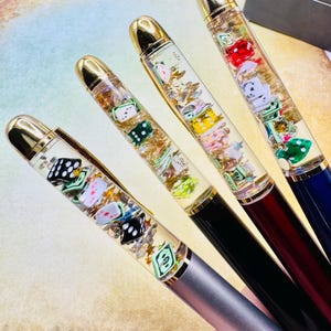 Puede incluir: Cuatro bolígrafos con barriles transparentes llenos de dados, dinero de juguete y copos dorados. Los bolígrafos tienen puntas y clips dorados, con barriles negros, plateados y burdeos. Los dados son de varios colores, incluyendo rojo, verde y blanco.