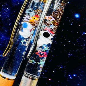 Op de afbeelding: Twee doorzichtige pennen met gouden en zilveren accenten, gevuld met kleurrijke sterren, astronautfiguren en kleine kralen. De pennen staan tegen een donkerblauwe achtergrond met een sterrenpatroon.