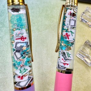 May include: Two pens with gold accents, one purple and one pink. The clear barrels contain floating Mahjong tiles, teal glitter, and gold flakes. The pens have a gold clip and a gold top.