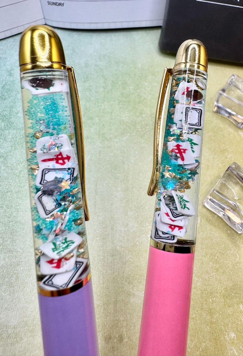 May include: Two pens with gold accents, one purple and one pink. The clear barrels contain floating Mahjong tiles, teal glitter, and gold flakes. The pens have a gold clip and a gold top.