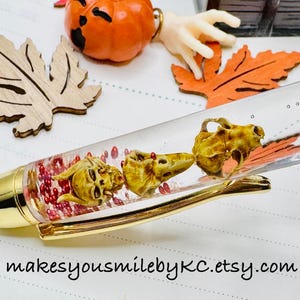 May include: A clear pen with a gold-coloured tip and accents, filled with small red beads and three miniature animal skulls. The pen is decorated with a pumpkin, wooden leaves, and the text "makesyousmilebyKC.etsy.com".