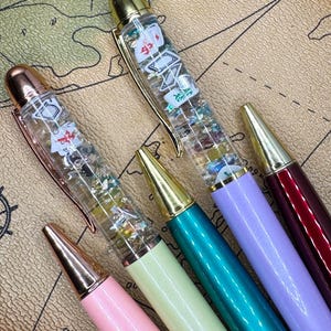 May include: A collection of colorful pens with gold-colored accents. The pens have clear barrels filled with glitter and small playing card-themed charms. The pens are in shades of pink, green, teal, purple, and burgundy.