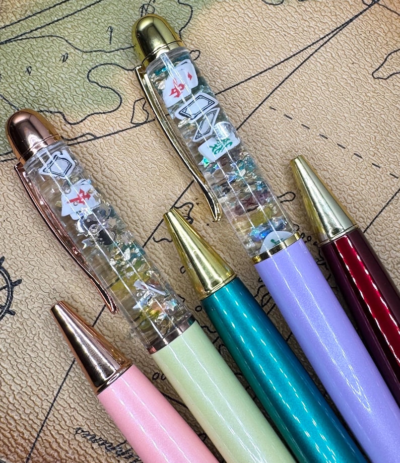 May include: A collection of colorful pens with gold-colored accents. The pens have clear barrels filled with glitter and small playing card-themed charms. The pens are in shades of pink, green, teal, purple, and burgundy.