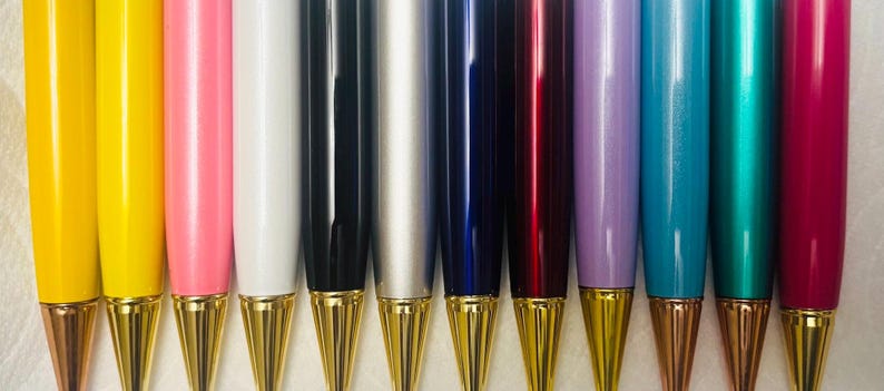 May include: A collection of colorful pens with gold-colored tips. The pens are arranged in a row, showcasing a variety of colors including yellow, pink, white, black, silver, blue, red, purple, teal, and magenta. The pens have a shiny finish.