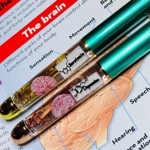 Puede incluir: Dos bol&iacute;grafos con tapas met&aacute;licas verdes y barriles transparentes. Cada barril contiene un cerebro en miniatura y las estructuras qu&iacute;micas de la serotonina y la dopamina. Los bol&iacute;grafos descansan sobre un diagrama del cerebro.