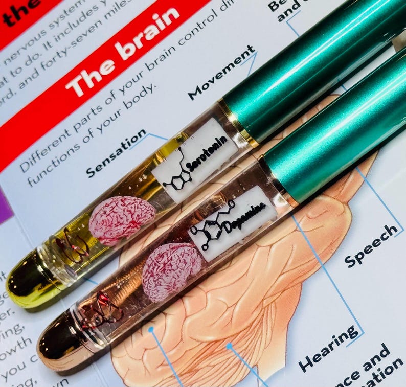 Puede incluir: Dos bol&iacute;grafos con tapas met&aacute;licas verdes y barriles transparentes. Cada barril contiene un cerebro en miniatura y las estructuras qu&iacute;micas de la serotonina y la dopamina. Los bol&iacute;grafos descansan sobre un diagrama del cerebro.