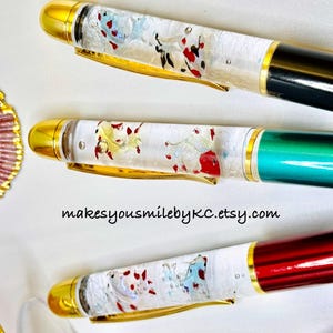 Puede incluir: Primer plano de tres bolígrafos coloridos con detalles dorados. Los bolígrafos tienen barriles transparentes llenos de elementos decorativos rojos, blancos y azules. Los capuchones de los bolígrafos son dorados y los cuerpos de los bolígrafos son negros, verdes y rojos. El texto "makesyousmilebyKC.etsy.com" es visible.