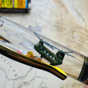 May include: A clear pen barrel containing a miniature olive green military helicopter model and small colorful star-shaped confetti. The pen features a gold-colored clip and a black grip section, showcasing a detailed rotor assembly inside.