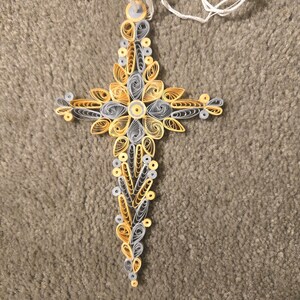 May include: A handcrafted cross pendant made from paper quilling. The cross features intricate designs in shades of yellow, gray, and white, with a white cord for hanging. The quilled paper is arranged in floral and geometric patterns.