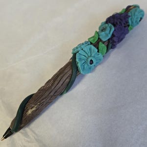 May include: A decorative pen with a brown, textured barrel, adorned with turquoise and purple flowers, green leaves, and a dark green vine. The pen has a black tip and a gold-colored point.
