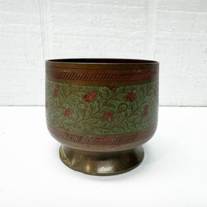 May include: A brass planter with a detailed floral design in green and red. The planter has a dark brown rim and base, and a pedestal base. The floral pattern is etched into the brass, creating a textured appearance.