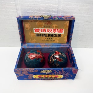May include: A blue box with a gold clasp contains two dark green iron balls with red and gold dragon designs. The box has a gold and red label and a printed card that reads "IRON BALL DIRECTION".
