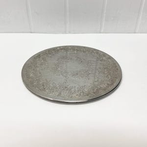 May include: A round silver-toned metal tray featuring an intricate embossed floral pattern along the outer rim and a smooth, reflective center. The tray has a low profile and a polished finish, showcasing detailed scrollwork and leaf motifs.