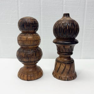 May include: Two dark brown wooden decorative objects with carved textures. The left object features three stacked spherical segments with a grid pattern. The right object has a rounded top with vertical carvings and a flared base with diagonal lines.