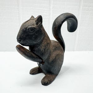 May include: A dark brown, textured cast iron squirrel figurine depicted in a seated position with its tail curved upward. The squirrel is holding a small, smooth, oval-shaped object near its chest, featuring a visible seam along the head and body.