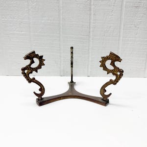 May include: A bronze-colored metal display stand with a decorative dragon design on each side. The stand has a curved base and a central support for holding an item. The dragon figures add an ornate touch to the piece.