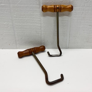 May include: Two vintage meat hooks with wooden handles. The handles are a light brown color, and the metal hooks are a dark, aged metal. One hook is lying down, and the other is standing upright.