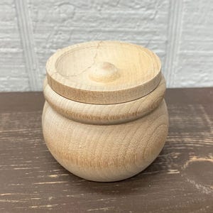 May include: A small, unfinished wooden jar with a lid. The jar is round with a slightly bulbous base and a lid with a small knob. The wood grain is visible, and the jar appears to be new. The jar is sitting on a brown surface.