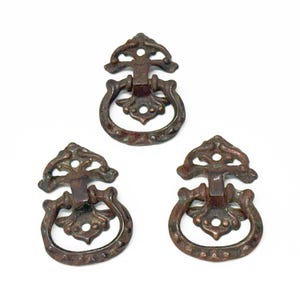 May include: Three antique bronze drawer pulls with ornate designs. Each pull has a ring handle and a decorative top.