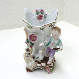 May include: A white porcelain vase with a boy figurine holding a bird and a goose figurine. The vase is decorated with pink and green flowers and gold accents.