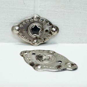 May include: Two antique silver-toned metal decorative pieces. Each piece has an oval shape with a star in the center and star and dot detailing around the edges. The pieces show signs of age and wear, with some areas of discoloration.