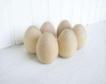 May include: Six wooden eggs in various sizes, arranged in a semi-circle on a white surface. The eggs are unpainted and have a natural wood finish.