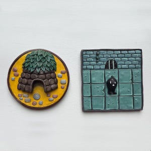 May include: Two decorative relief sculptures. One is a brown and green beehive shape on a yellow and brown circular base. The other is a teal square with a dungeon scene and a black figure.