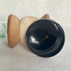 Puede incluir: Un botón negro brillante con un pequeño agujero está junto a una bobina de madera con hilo beige. El botón es redondo y ligeramente curvado. La bobina es de madera de color marrón claro.