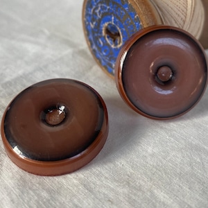 Puede incluir: Dos objetos marrones en forma de rosquilla con un acabado brillante. Los objetos tienen un centro elevado y un anillo marrón más oscuro alrededor del borde. Una bobina de madera con detalles azules y blancos está en el fondo. Los objetos están sobre una superficie blanca.