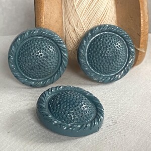 May include: Three teal buttons with a textured centre and a rope-like border. The buttons are round and slightly domed. The background includes a wooden spool and beige thread.