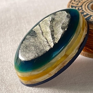 Op de afbeelding: Een ovaalvormige hars hanger met lagen blauw, geel en wit. De bovenste laag heeft een zilverkleurig, getextureerd ontwerp. De hanger heeft een donkerblauwe rand en ligt op een zachte, witte achtergrond.