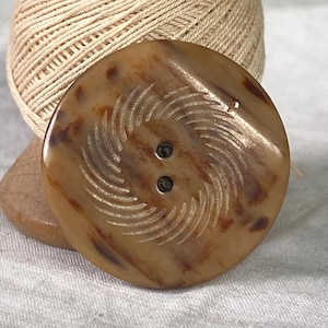May include: A large, round button with a swirl design carved into its surface. The button is a mottled brown color with two small holes for sewing. A spool of beige thread is in the background.