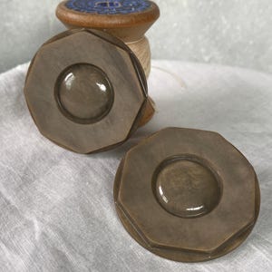 May include: Two brown, octagonal buttons with a circular center are displayed on a white cloth. A wooden spool with blue and brown details sits in the background. The buttons have a glossy finish.
