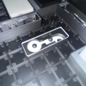 May include: A rectangular, black and white device with two circular white components and a black center. The device has a black border and two black screws. The background is a grid of gray squares.