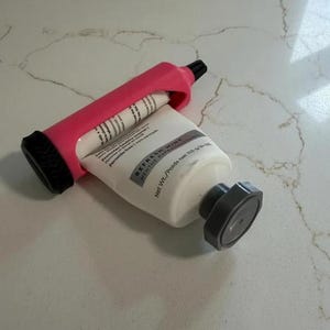 Puede incluir: Un tubo blanco de pasta dental Refresh Mint con tapa negra está siendo exprimido por un exprimidor de tubos rosa. El exprimidor tiene un rodillo negro y está diseñado para aprovechar al máximo el tubo de pasta de dientes.