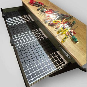 May include: A Husky brand workbench with a light wood top and an open drawer featuring a black and white grid-patterned liner. A collection of various hand tools, including screwdrivers, pliers, and wrenches, is scattered across the wooden surface.