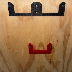 May include: Two metal brackets, one black and one red, mounted on a wooden surface. The black bracket is larger, with a rectangular design and two screw holes. The red bracket is smaller, with a U-shaped design. Both are secured with screws.