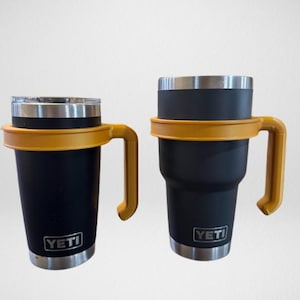 May include: Two black insulated tumblers with silver rims and yellow handles. The tumblers have the brand name "YETI" printed on the front. One is shorter and wider than the other.