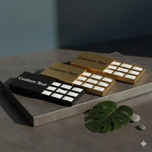 May include: Four rectangular boxes, two gold and two black, with the text "Custom Text" on the lids. Each box is open, revealing a grid of small compartments with white padding. The boxes are arranged on a gray surface.
