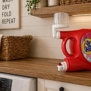 May include: A red Tide laundry detergent bottle with a white dispenser attached. The bottle has a floral design and the Tide logo. The image is set in a laundry room with a basket and a sign that says "Wash, Dry, Fold, Repeat."