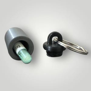 May include: A gray metal pill holder with a black cap and key ring. A green and white capsule is partially visible inside the holder. The capsule has the text "6mg T" printed on it. The cap is detached, showing the key ring.