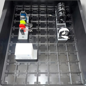 May include: A black storage container with a grid pattern, holding various tech accessories. Items include USB drives, charging cables, and a white box. The accessories are organized in separate compartments within the container.