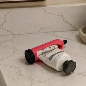 May include: A pink and black tube squeezer is squeezing a white tube with black text. The squeezer is on a white countertop with a marble pattern. A silver faucet and a soap dispenser are also visible.