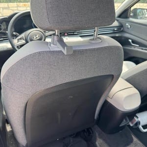 May include: Interior view of a car's back seat. The seat is upholstered in gray fabric with a light gray headrest. A small, gray hook is attached to the headrest. The car's interior is visible, including the steering wheel and center console.