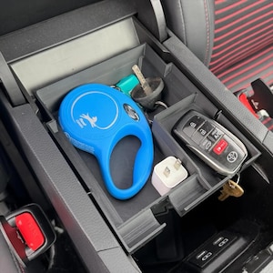 May include: A black car console organizer with multiple compartments. A blue retractable dog leash, car keys, a phone charger, and a car key fob are visible. The organizer is designed to fit inside a car's center console.