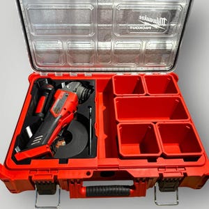 May include: A red and black Milwaukee Packout storage case with an open transparent lid. The interior features a molded foam insert holding a red angle grinder and a metal wrench, alongside four empty red plastic storage bins for organization.