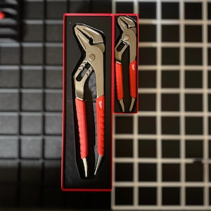 May include: Two metal pliers with red handles are displayed in a black box with a red border. The pliers have a silver finish and a textured grip. The box is on a dark surface.