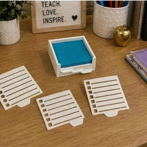 May include: A white desk organizer with a teal notepad and three white paper cutouts. A sign that says "TEACH. LOVE. INSPIRE." is in the background. A gold apple and a purple notebook are also on the desk.